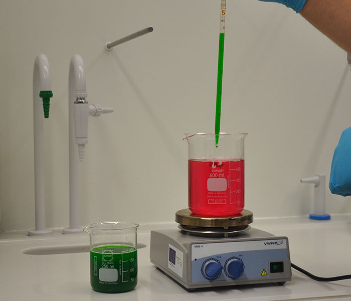 Punaista nestettä täynnä olevaan dekantterilasiin pipetoidaan vihreää nestettä. Dekantterilasi on magneettisekoittajan päällä. Sen vieressä on vihreää nestettä täynnä oleva dekantterilasi.