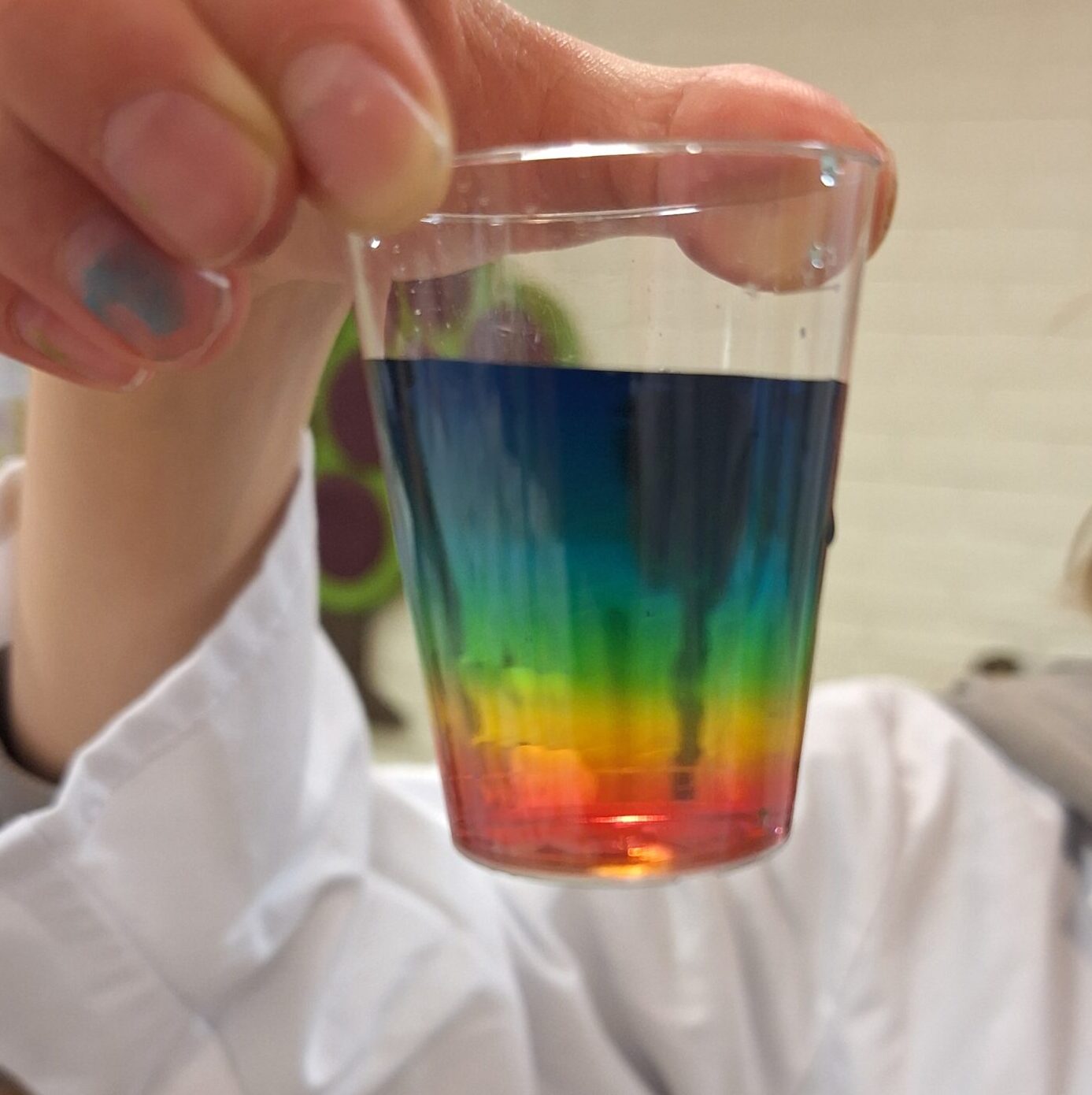 Muovinen snapsilasi, jonka sisällä on sateenkaaren värinen liuos.